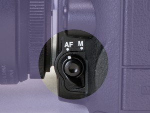 写真02フォーカス切り替え SW（ Nikon D7000 ） カメラの本体には、フォーカスモードを AF と M（ マニュアル ）に切り替えるスイッチが付いている。