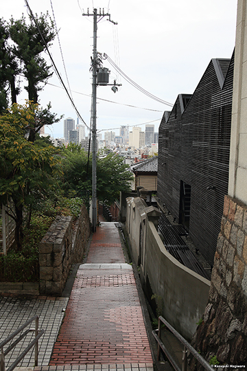 石畳の小径から、脇の石段の続く坂道を下る。坂の向こうには神戸の街並み。こうした光景、坂のある街ならではで、僕は好きだ　　Canon EOS 5D MarkIII ＋ Tokina AT-X 24-70 F2.8 PRO FX　　35mm　絞り優先 AE（ f9.0　1/250 秒 ）　ISO：800　AWB　RAW